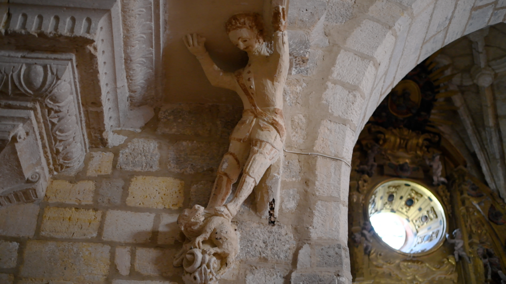 Ménsula barroca del coro que representa a un hombre vestido con casaca y calzas sosteniendo el borde del coro a la vez que pisa a un pequeño dragón que vuelve la cabeza hacia él.